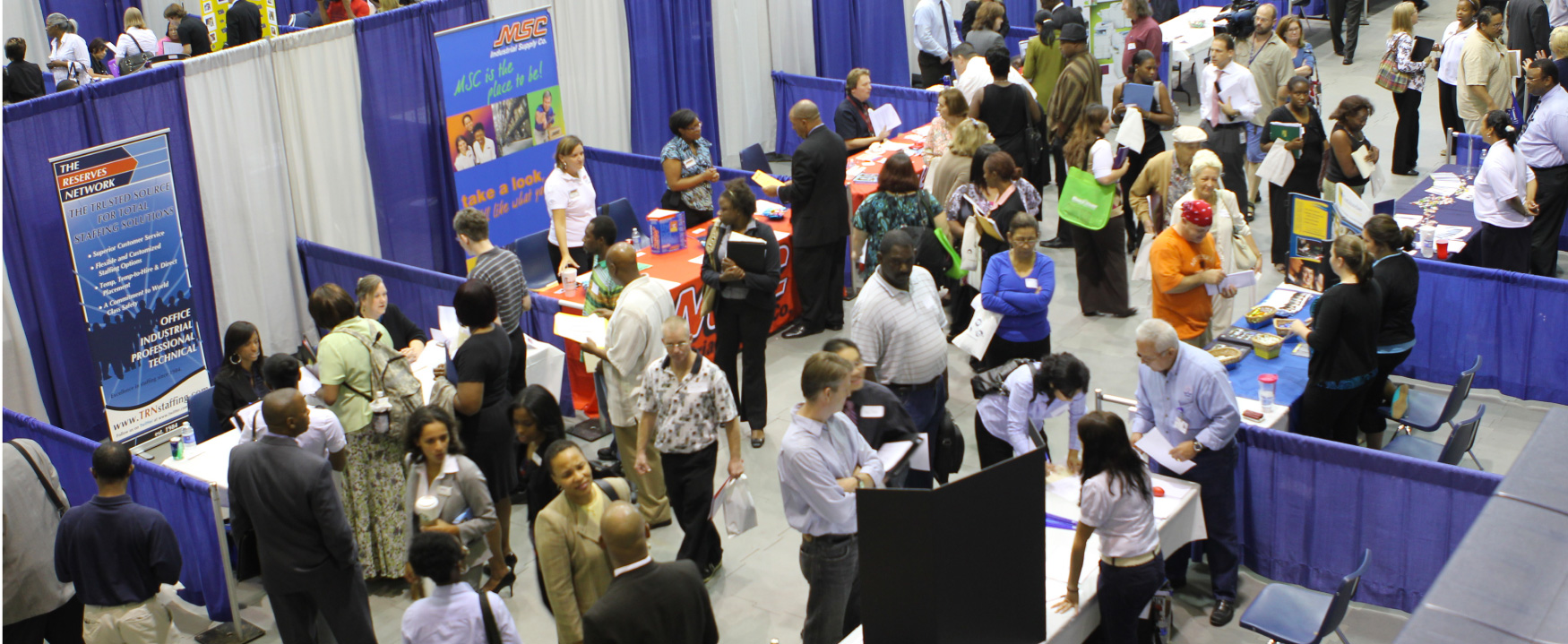 Inside a local civic center, there are rows of tables, with large advertisements behind each table. The space is filled with people walking around, visiting the people standing behind each table.