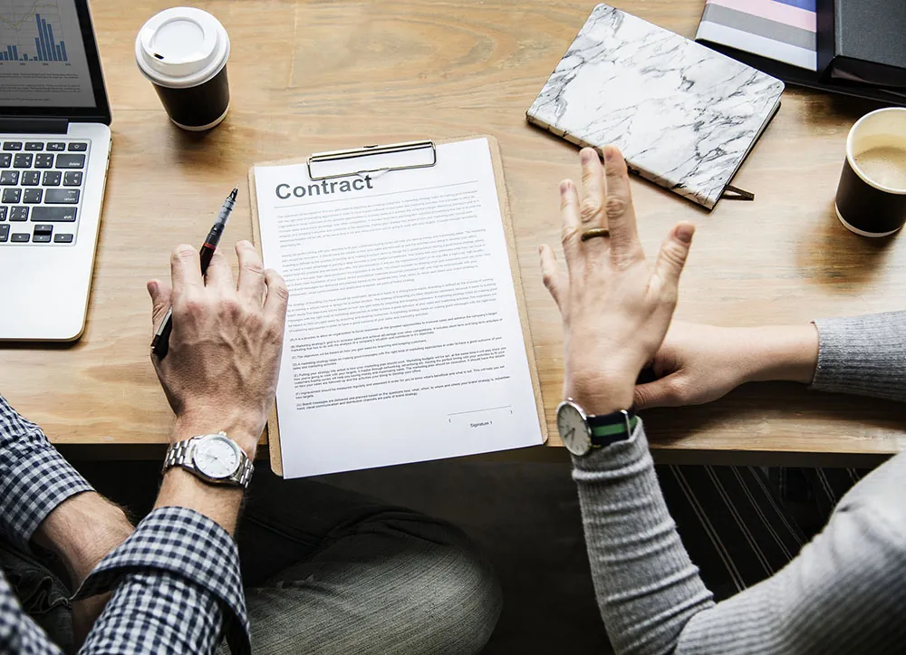 Two people sitting at a table with their hands above a contract that is on a clipboard on the table.