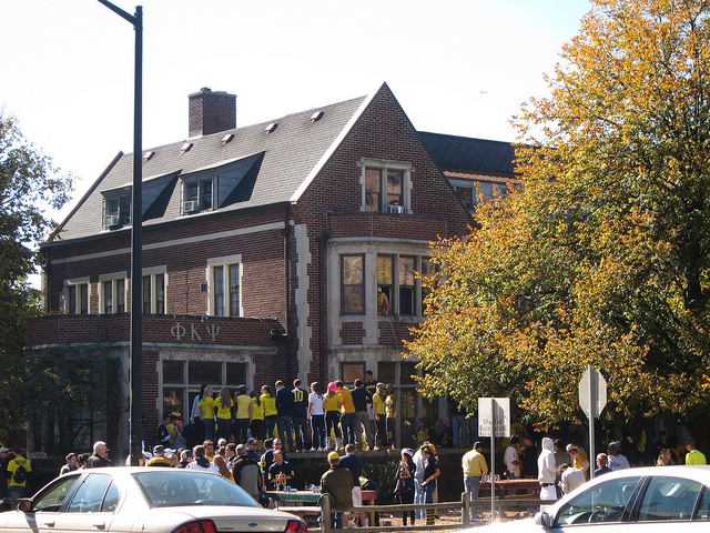 "A large group of people, many in yellow shirts, gathered outside a brick building with the Greek letters Phi Kappa Psi (ΦΚΨ) on the front. The scene includes autumn trees and several parked cars in the foreground."