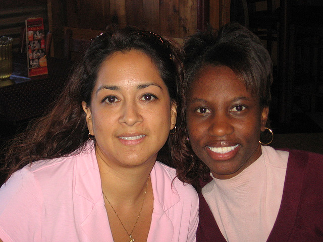 "Two people sitting closely together in a dimly lit indoor setting. The person on the left has long, wavy hair and is wearing a light pink top with a gold necklace. The person on the right has short hair and is wearing a maroon top over a light-colored shirt. Wooden paneling, chairs, and tables in the background suggest a restaurant or café setting. Both people are people of color."
