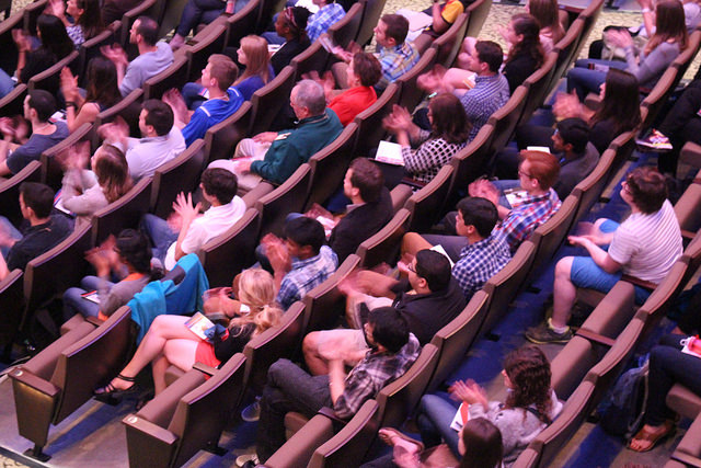 "A group of people seated in tiered rows of chairs, likely in an auditorium or lecture hall. Many are clapping, suggesting a response to a performance or presentation. Some individuals have notebooks or papers on their laps."