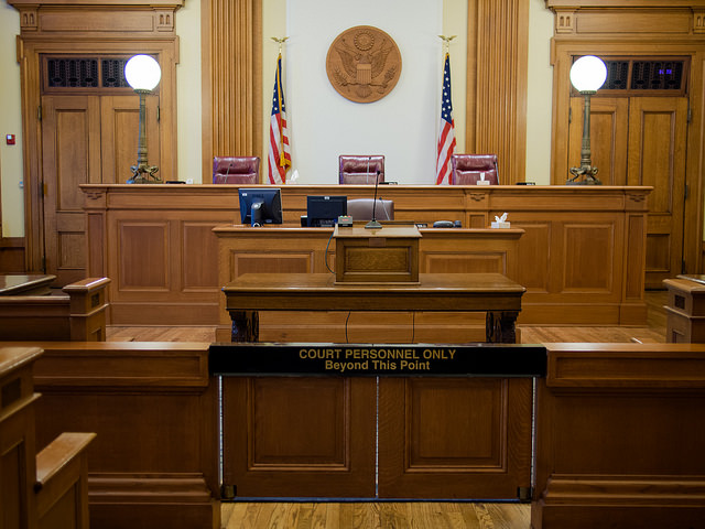 A courtroom with wooden furnishings, featuring a judge's bench at the front with three chairs behind it. The United States seal is mounted on the wall above the bench, flanked by two American flags. There are two computer monitors and other office supplies on the bench. In front of the judge's bench is a barrier with a sign that reads "COURT PERSONNEL ONLY Beyond This Point." The courtroom has an orderly and formal appearance, typical of judicial settings.