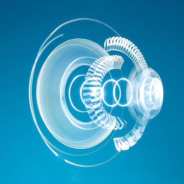 The image shows a complex light painting against a blue background. The light painting consists of multiple concentric circles and arcs, with varying thicknesses and brightness. Some parts of the light painting resemble gears or mechanical components, creating an intricate and abstract design. The overall effect is dynamic and visually captivating, with the bright white lights contrasting sharply against the deep blue backdrop