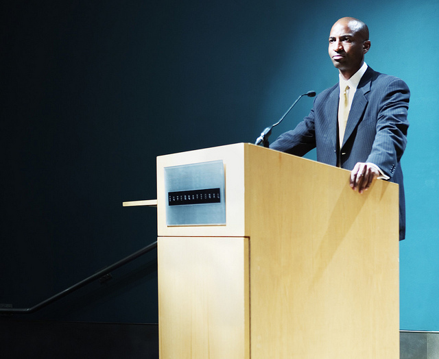 "A person in a suit stands behind a wooden podium with a microphone, delivering a speech or presentation."