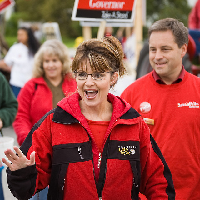 "A group of people participate in a political campaign event. The central figure, wearing a red and black 'Mountain Hard Wear' jacket, gestures with one hand. In the background, others hold signs that read 'Governor' and 'Sarah Palin.'"