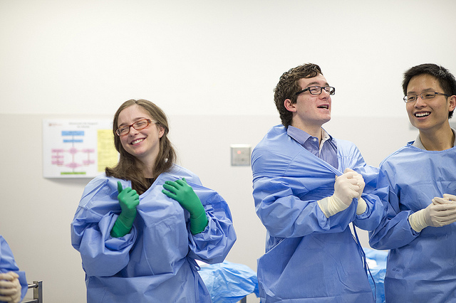 "Three individuals in a medical setting wear blue surgical gowns and gloves. The person on the left has green gloves, while the other two wear white gloves. A poster with text and diagrams is visible on the wall in the background."
