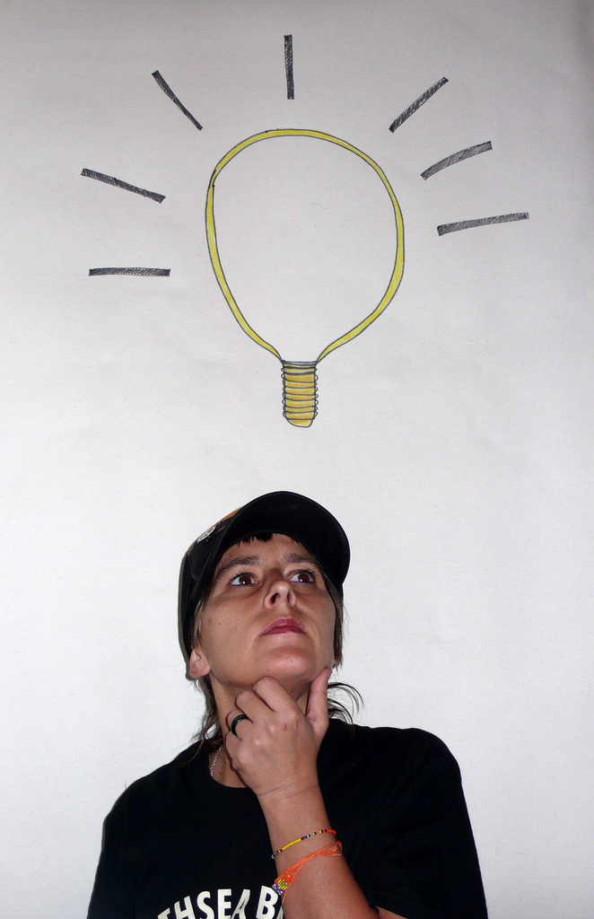 "A person in a black shirt and black cap stands against a white background. Above their head is an illustrated yellow light bulb with black rays, symbolizing an idea or inspiration."