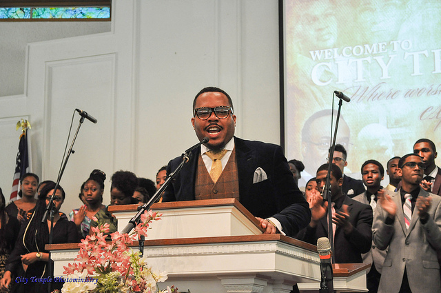 "A person speaks at a podium in a church setting, with several individuals standing behind them—some holding microphones. The podium is decorated with flowers. In the background, a large screen displays the text 'WELCOME TO CITY TEMPLE' along with images of people. An American flag is visible on the left side of the image."