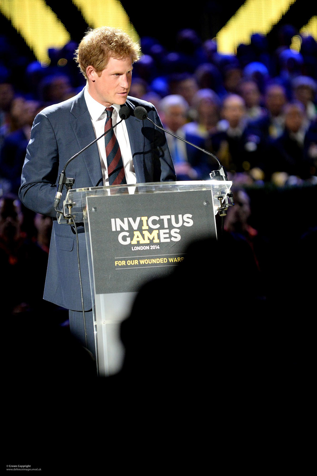 "A person in a suit and tie stands at a podium with a microphone, delivering a speech. The podium displays a sign that reads 'INVICTUS GAMES LONDON 2014 FOR OUR WOUNDED WARRIORS.' In the background, an audience with blurred faces is visible, and bright yellow lights form an 'X' pattern above the crowd."