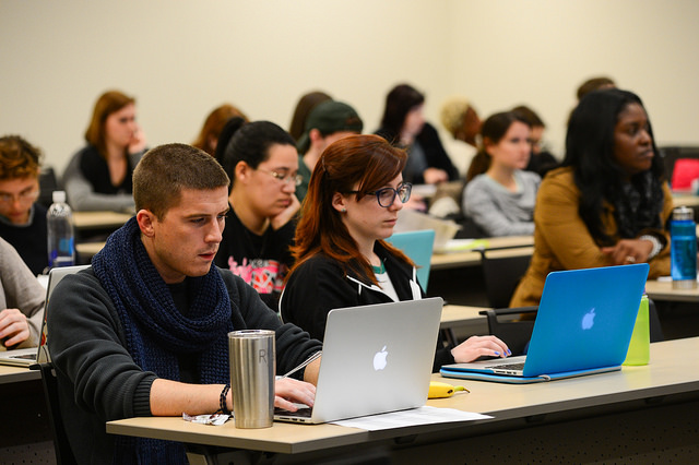 "A classroom filled with students sitting at desks, each working on a laptop. Various laptops, including several Apple MacBooks, are visible. Some desks have water bottles and coffee mugs. The classroom has beige walls and a whiteboard at the front."