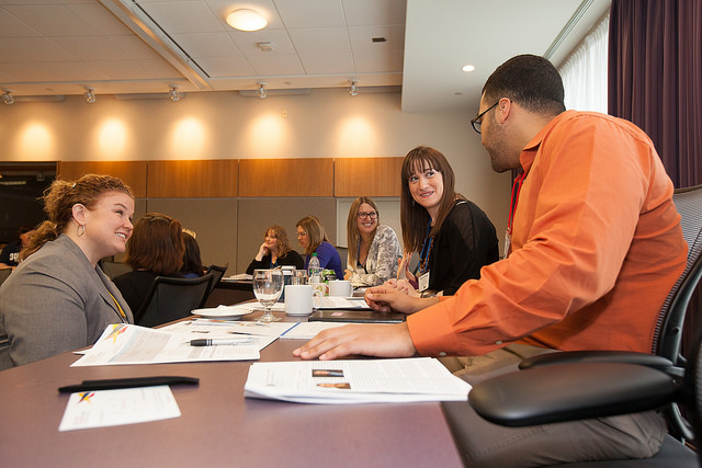 "A group of people are seated around a conference table in a well-lit meeting room. The table is covered with papers, notebooks, and glasses of water. Some individuals are taking notes while others are engaged in conversation. The background features wood paneling and a large window with curtains."