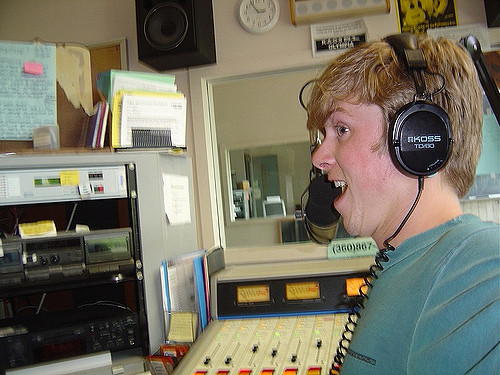 "A person wearing headphones stands in a radio station control room, facing away from the camera. The room contains a soundboard with numerous buttons and sliders, audio equipment on shelves, and a bulletin board with pinned papers. Books and folders are also visible on top of the shelves."