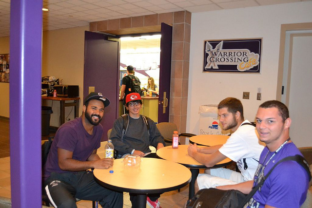 "Four individuals are seated around a circular table in a casual indoor setting. The table holds several items, including a bottle of water and other drinks. In the background, an open door leads to another room where more people are visible. A sign on the wall reads 'Warrior Crossing Cafe.'"