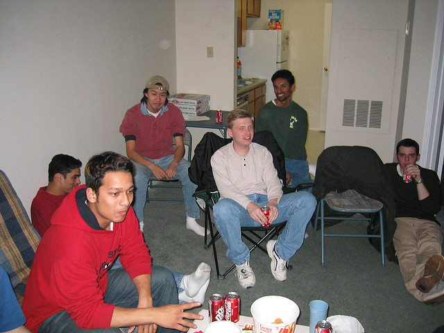 "A group of six people are sitting in a living room on chairs and a couch, facing the camera. A table in front of them holds several cans of soda and snacks, including a bowl of chips. In the background, an open door reveals a kitchen with more food items on the counter."