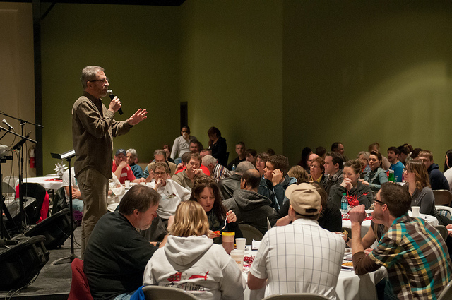 "A man stands on a stage holding a microphone and speaking to an audience seated at round tables. The audience includes many people, some watching the speaker and others conversing. The room has green walls, is well-lit, and features musical instruments and equipment on the left side of the stage."