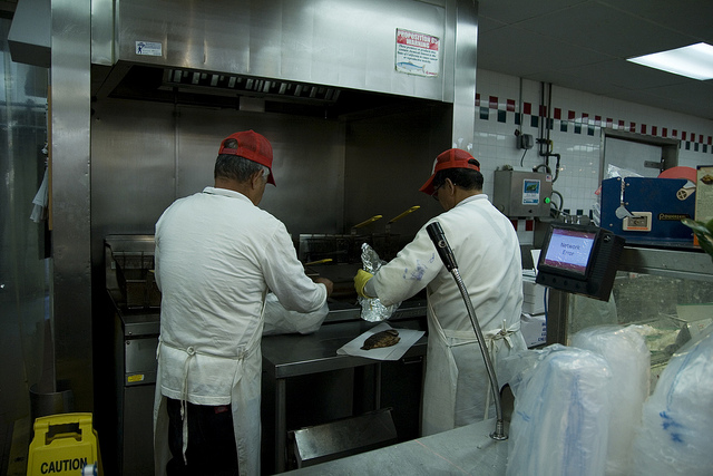 "Two workers in white uniforms and red caps prepare food in a commercial kitchen. They stand at a stainless steel counter with cooking equipment, including a fryer, in the background. One worker uses tongs to handle foil-wrapped food, while the other places food on a piece of paper. A caution sign is on the floor to the left, and a monitor displaying 'Order Ready' is visible on the right."