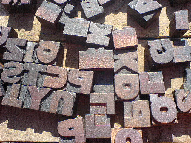 "A collection of wooden letterpress blocks is scattered on a wooden surface. The blocks display various letters and numbers, including 'P,' 'S,' 'T,' 'D,' 'K,' 'L,' and '9,' arranged haphazardly with some facing different directions. The wood appears worn, suggesting frequent use."