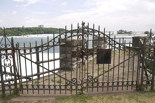 "An ornate wrought iron gate with intricate designs stands closed, blocking access to a viewing area overlooking a large body of water. The gate is flanked by stone pillars, with a small plaque mounted on the right. Rapids or waterfalls are visible in the background, along with distant buildings under a partly cloudy sky."