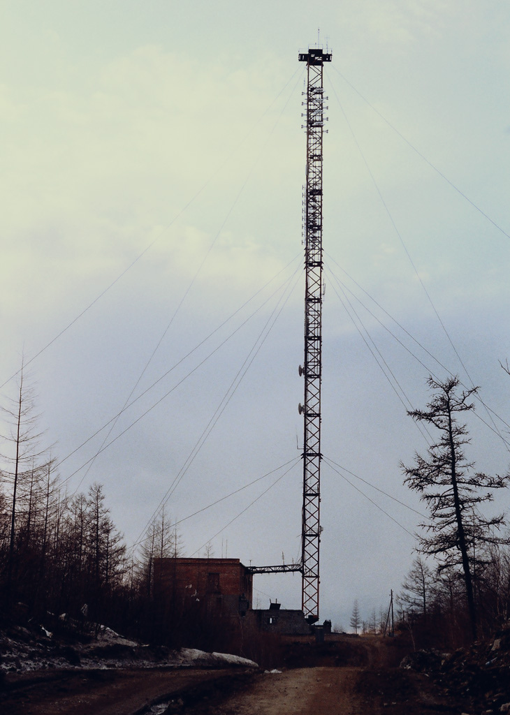 "A tall metal communication tower stands in a remote area with sparse trees, supported by multiple guy wires. At its base is a small brick building with an antenna on the roof. The sky is overcast with light clouds, and a dirt road leads to the building and tower, with patches of snow along the roadside."