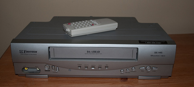 "An Emerson VCR labeled 'DA-4HEAD' with a remote control resting on top. The front panel features buttons for power, eject, play, stop, rewind, fast forward, and record, along with audio/video input and output ports. Specifications like 'Hi-Fi HQ' and '19 Micron Heads' are visible. The device sits on a wooden surface against a plain background."