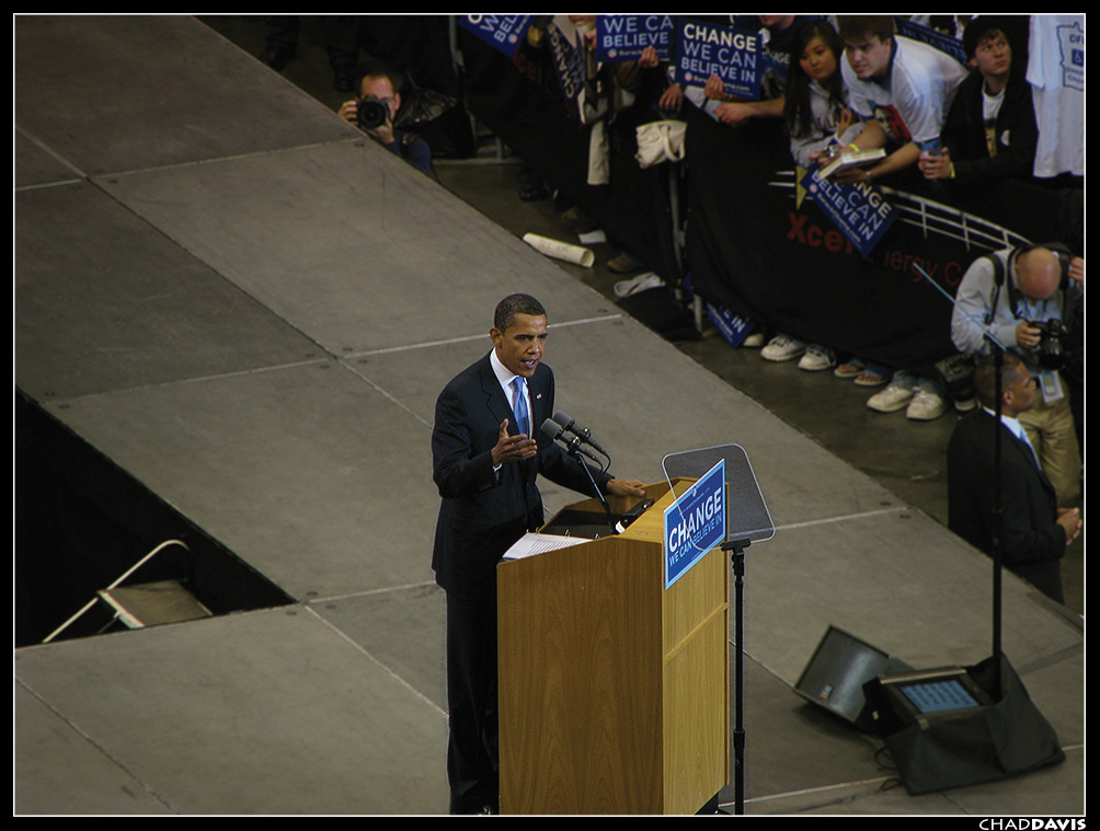 "Barack Obama in a suit stands at a podium giving a speech. The podium displays a sign that reads 'CHANGE WE CAN BELIEVE IN' and is equipped with multiple microphones. Behind him, an audience holds signs with similar messages, and photographers are taking pictures. The background includes part of an elevated stage and some equipment on the floor."