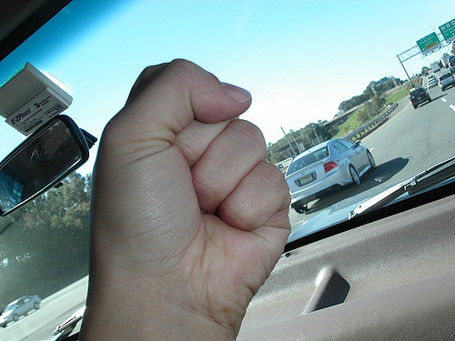 A close-up of a person's hand making a fist inside a car. The photo is taken from the driver's perspective, showing the dashboard and part of the windshield. Outside, there is a white car driving on the highway with other vehicles in the background. The sky is clear and blue.