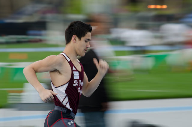"A runner in a maroon and white athletic uniform is captured mid-stride during a race. The background is blurred, indicating motion, with green grass and other indistinct figures visible. The runner's face is pixelated for anonymity."