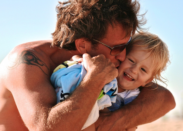 "A shirtless person with a tattoo on the upper arm is holding a small child wrapped in a towel. Both faces are blurred out. The background shows a clear blue sky, suggesting they are outdoors, possibly at the beach."