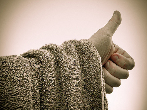 "A close-up image of a person's hand giving a thumbs-up gesture. The person is wearing a textured, fuzzy robe or towel that covers the arm up to the wrist. The background is plain and out of focus, emphasizing the hand and thumb."