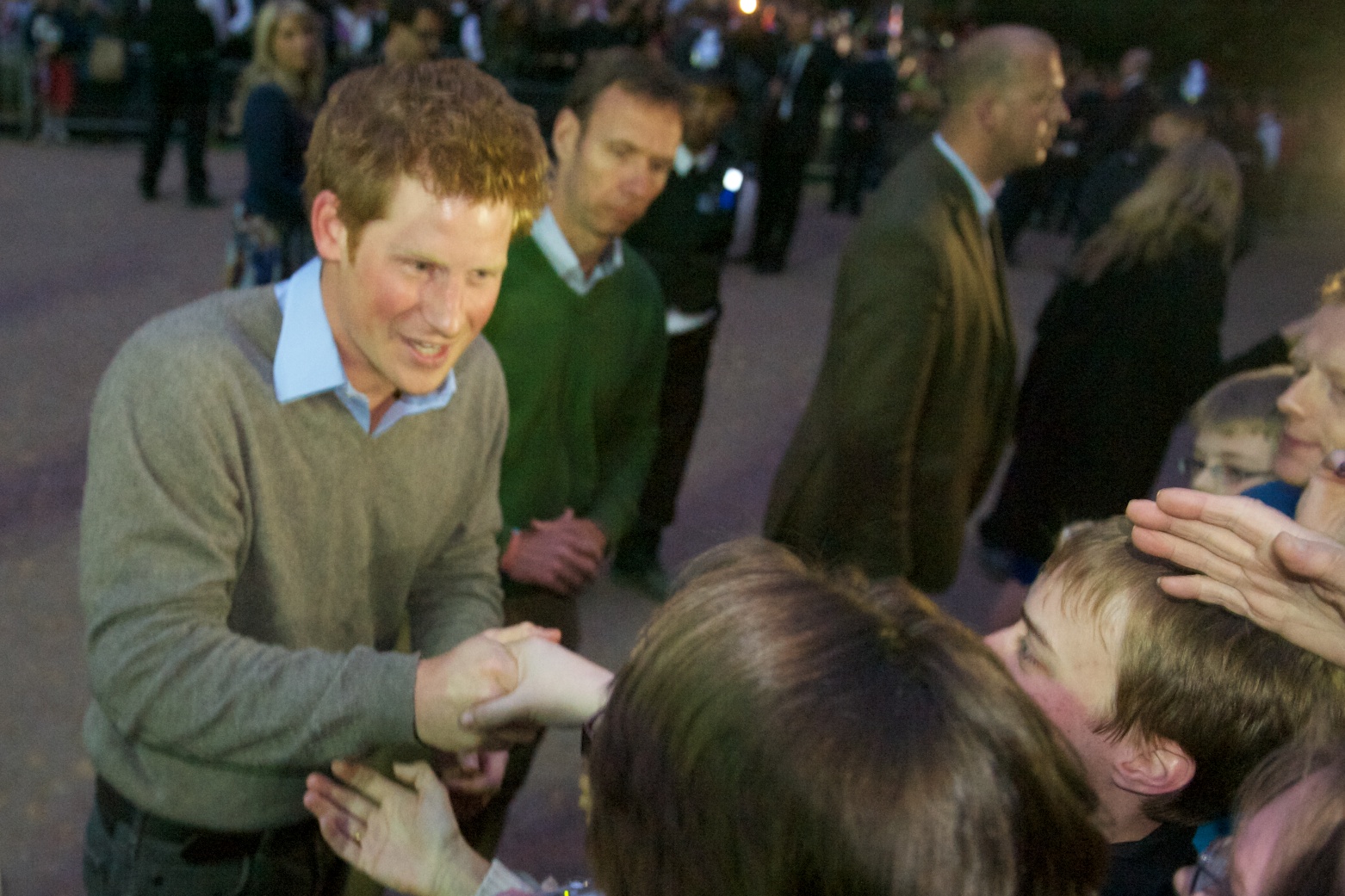 "A man with curly hair, wearing a gray sweater over a light blue collared shirt, is shaking hands with a person in the foreground. Several other people are visible in the background, some of whom are also interacting with others. The image appears to be taken at an outdoor event during the evening. Faces of all individuals have been blurred for privacy."