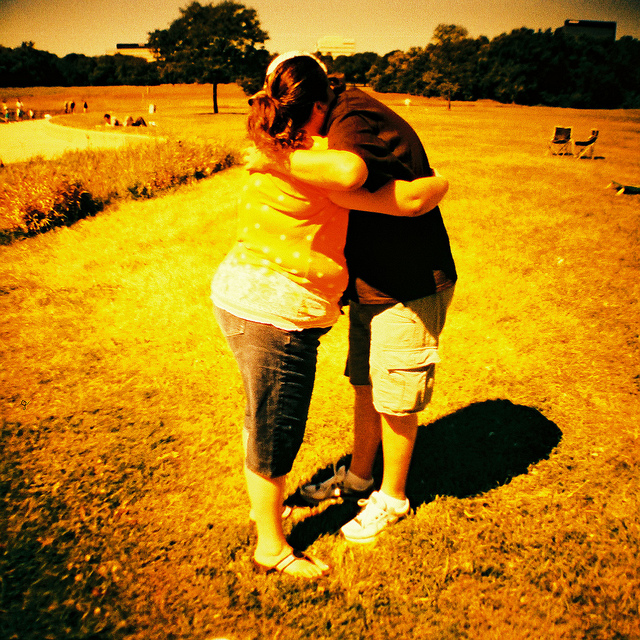 "Two people are hugging in a grassy field on a sunny day. The person on the left is wearing an orange sleeveless top and blue jeans, while the person on the right is wearing a black t-shirt and beige shorts. There are trees and other people in the background, as well as two empty chairs to the right. The image has a warm, yellowish tint."