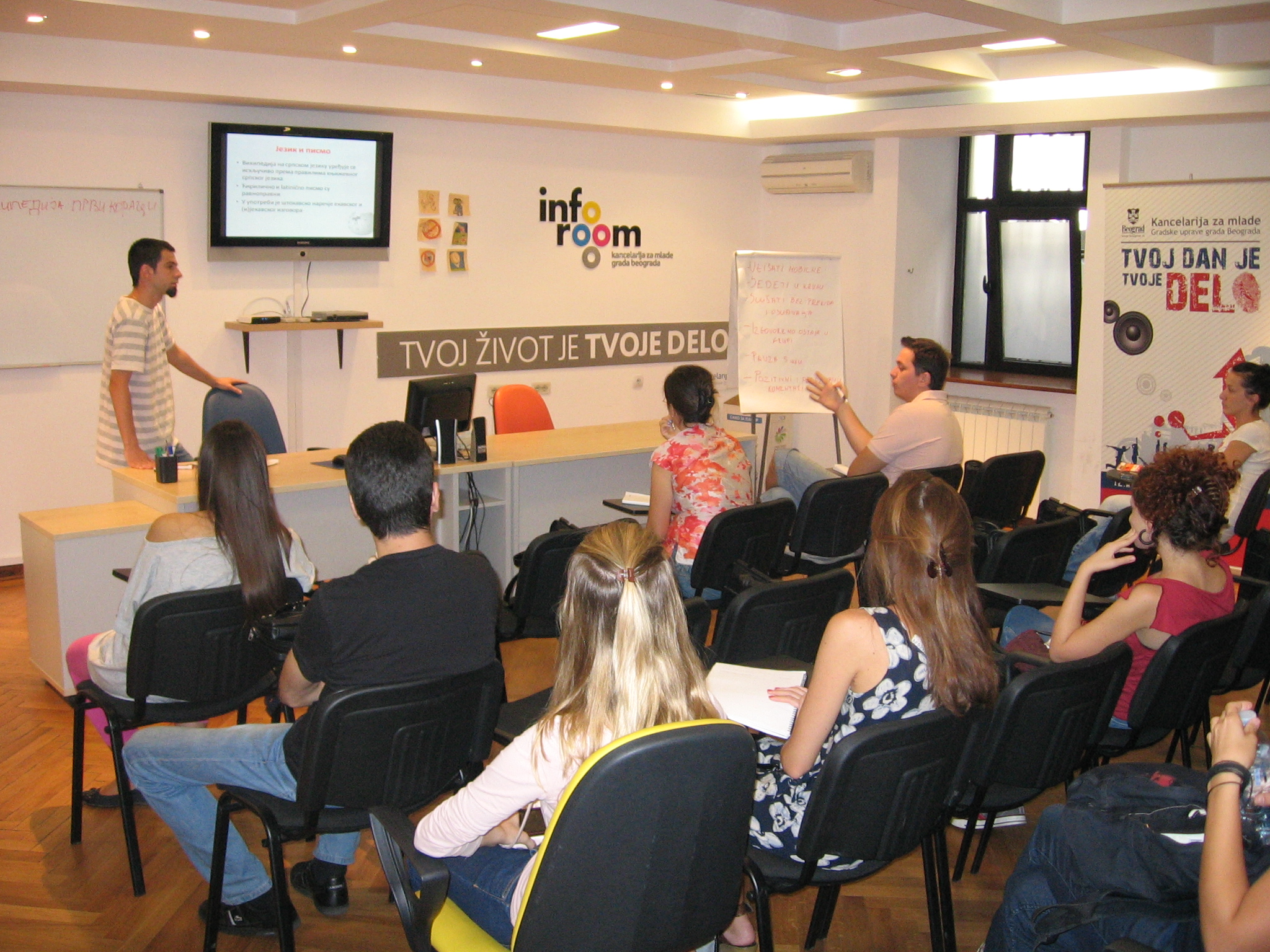 "A classroom setting with a group of students seated and facing a presenter at the front. The presenter is standing next to a large screen displaying a presentation slide. There are various posters and banners on the walls, including one that reads 'info room' and another that says 'TVOJ ŽIVOT JE TVOJE DELO.' One student is writing on a whiteboard while others are attentively listening. The room has wooden flooring and modern lighting." This image captures an educational or training session taking place in a classroom environment, highlighting an interactive learning experience with visual aids, active participation from students, and motivational messages displayed around the room.