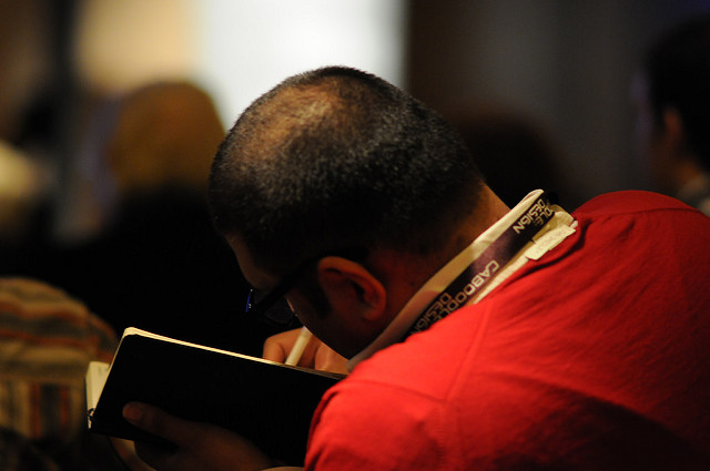 "A person with short hair and glasses is writing in a notebook. The person is wearing a red shirt and a lanyard around the neck. The background is blurred, focusing on the individual who appears to be taking notes or sketching something in the notebook."