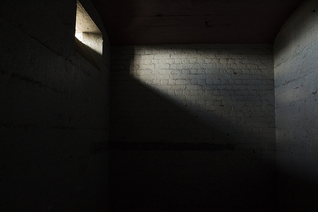 Here’s the alt text for the image: "A dimly lit room with a small rectangular window near the top left corner. Light from the window casts a diagonal beam across a rough, white brick wall, creating a stark contrast between light and shadow. The rest of the room remains in darkness."
