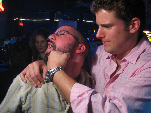 "Two people are standing close together in a dimly lit setting, possibly a bar or nightclub. One person, wearing a light-colored striped shirt, has an arm around the other’s shoulder. The second person, in a pink checkered shirt, has their hand on the first person’s face. Neon lights and other people are visible in the background."