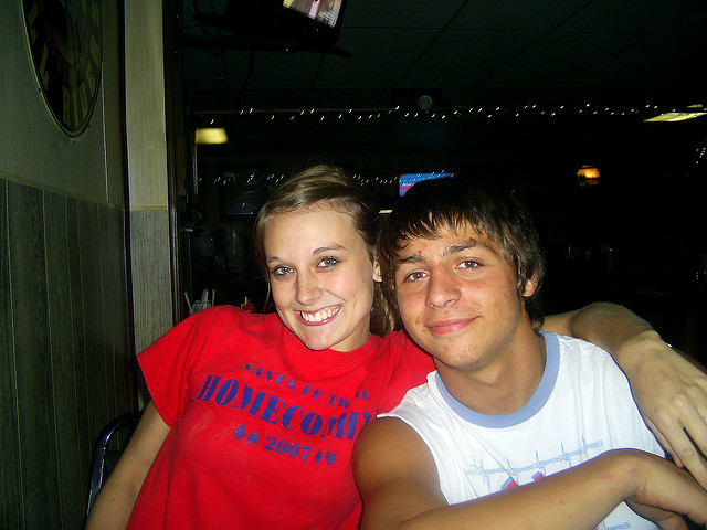 "Two people are sitting closely together in a dimly lit room. The person on the left is wearing a red t-shirt with blue text that reads 'Homecoming 2007,' and the person on the right is in a white sleeveless shirt with a design. String lights hang from the ceiling in the background, with other indistinct objects and people visible in the dim lighting."