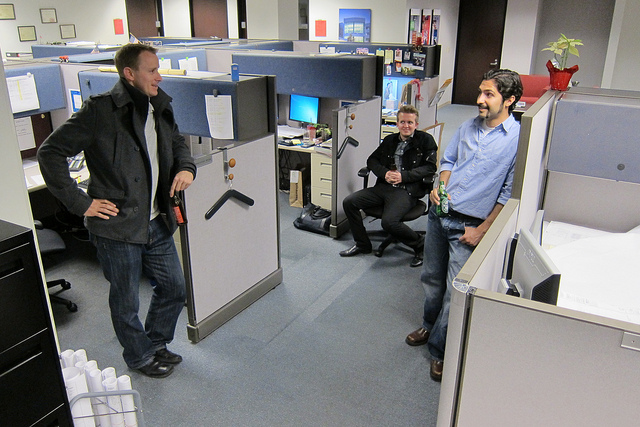 "An office environment with three people in a cubicle area. Two are standing and one is seated on a chair. The cubicles contain office supplies, papers, and personal items. One person is holding a drink, another has a jacket draped over an arm, and a small potted plant sits atop one of the cubicles."