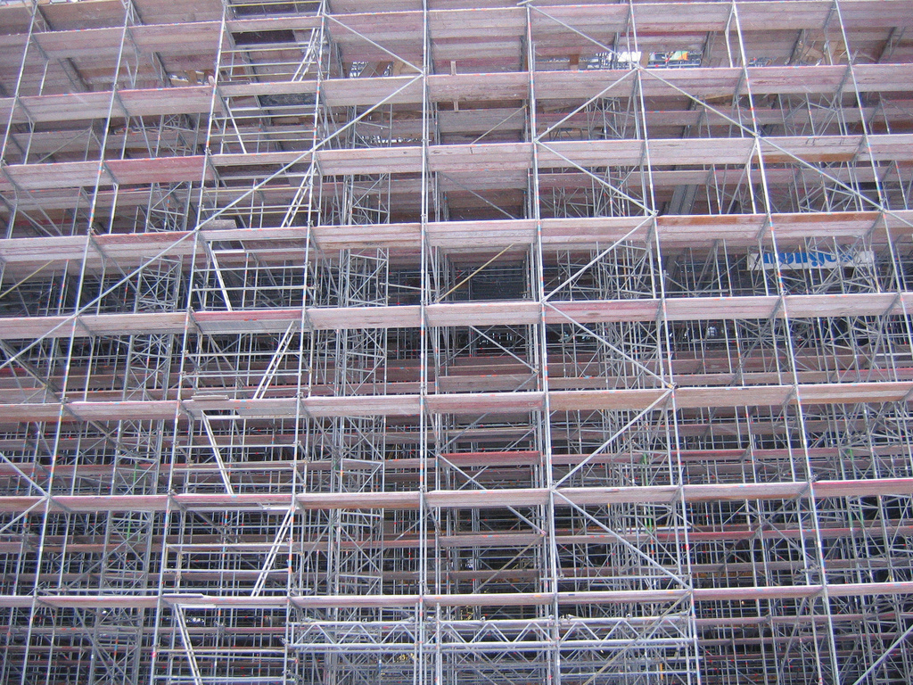 "A large, complex structure of metal scaffolding with multiple levels and cross-bracing, forming a grid-like pattern. The scaffolding appears to be part of a construction site or building renovation project."