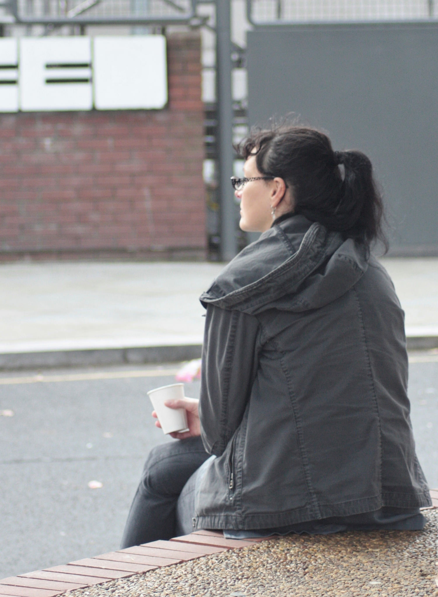 A person with dark hair tied in a ponytail is sitting on a ledge outdoors, wearing a gray jacket and holding a white cup. The background includes a brick wall and some mailboxes. The image captures a moment of solitude or contemplation in an urban setting, with the person's face blurred for privacy, emphasizing the scene rather than the individual's identity.