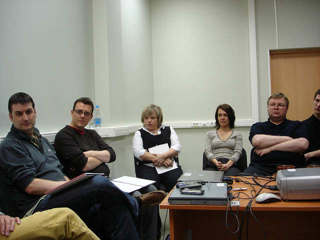 A group of six people are seated in a semi-circle in a small, plain room. They appear to be engaged in a meeting or discussion. There is a table in the foreground with various items on it, including a laptop, papers, and cables.