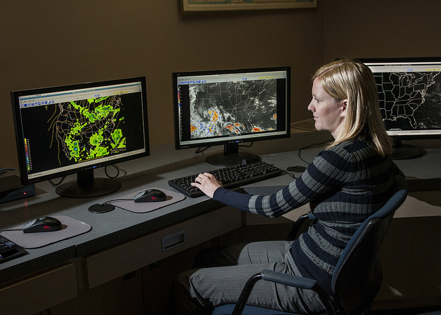 A person is seated at a desk with three computer monitors displaying weather maps. The left monitor shows a map with green radar data indicating precipitation. The middle monitor displays a satellite image with cloud formations and some colored areas representing different weather conditions. The right monitor shows another weather map, likely focusing on a different region or aspect of the weather. The image captures a person analyzing weather data on multiple computer screens, illustrating how meteorologists or weather analysts work to predict and track weather patterns.
