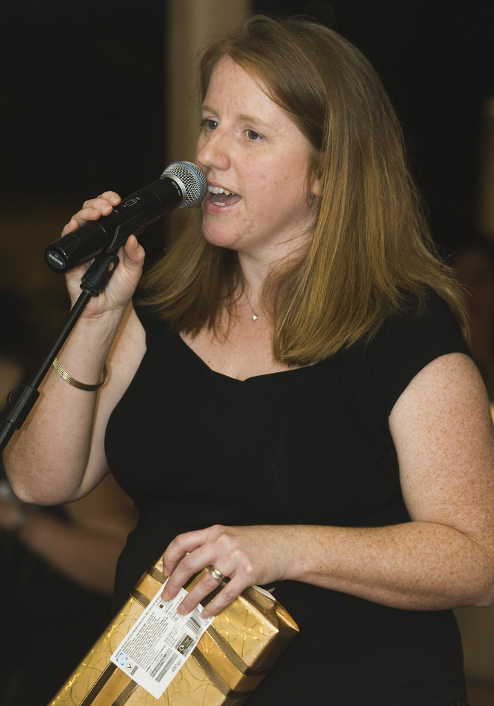 "A person with shoulder-length hair, wearing a black top, holds a microphone in one hand and a wrapped gift in the other. The background is blurred and indistinct."