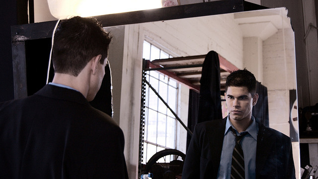 "A man in a suit and tie looks at himself in a mirror. His reflection in the mirror is shown. The background features a large window with natural light, an industrial-style shelf, and some hanging clothes."