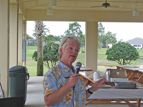 "A person stands under a pavilion, holding a microphone and speaking. They are wearing a blue shirt with yellow floral patterns. In the background, picnic tables hold trays and containers, and the pavilion is set in an outdoor area with green grass, trees, and distant buildings."