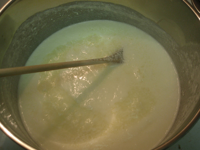 "A close-up of a pot containing a thick, creamy white mixture being stirred with a wooden spoon. The mixture appears to be in the process of cooking or preparation."