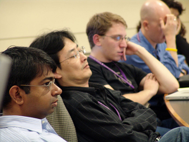 "A group of people sit in a row facing forward, likely attending a meeting or lecture. Some wear lanyards, suggesting a conference or seminar setting. The indoor background features beige walls and a table in front of them."