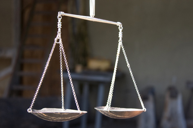 "A close-up of a traditional balance scale with two metal pans suspended by chains on either side of a horizontal beam. The background is blurred, showing an indoor setting with indistinct objects and furniture."