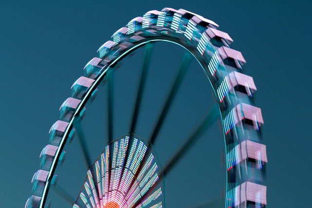 "A long-exposure photograph of a Ferris wheel at night, brightly lit with colorful lights that create a blurred motion effect as it spins. The dark sky in the background highlights the vibrant illumination."