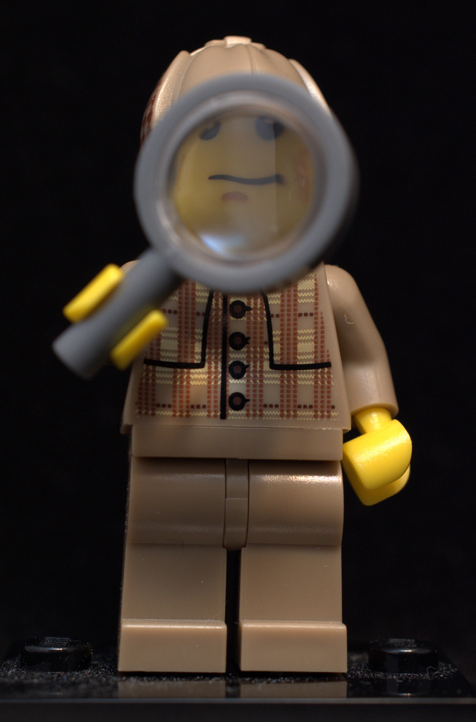 "A Lego minifigure dressed as a detective holds a magnifying glass up to its face. The figure wears a brown plaid jacket and brown pants, with yellow hands and head, set against a black background."