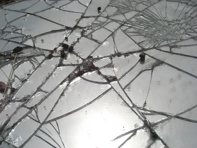"A close-up view of a shattered glass surface with numerous cracks and broken pieces. Light reflects off the glass, creating a bright spot in the center. Small debris and dirt particles are visible on the surface."